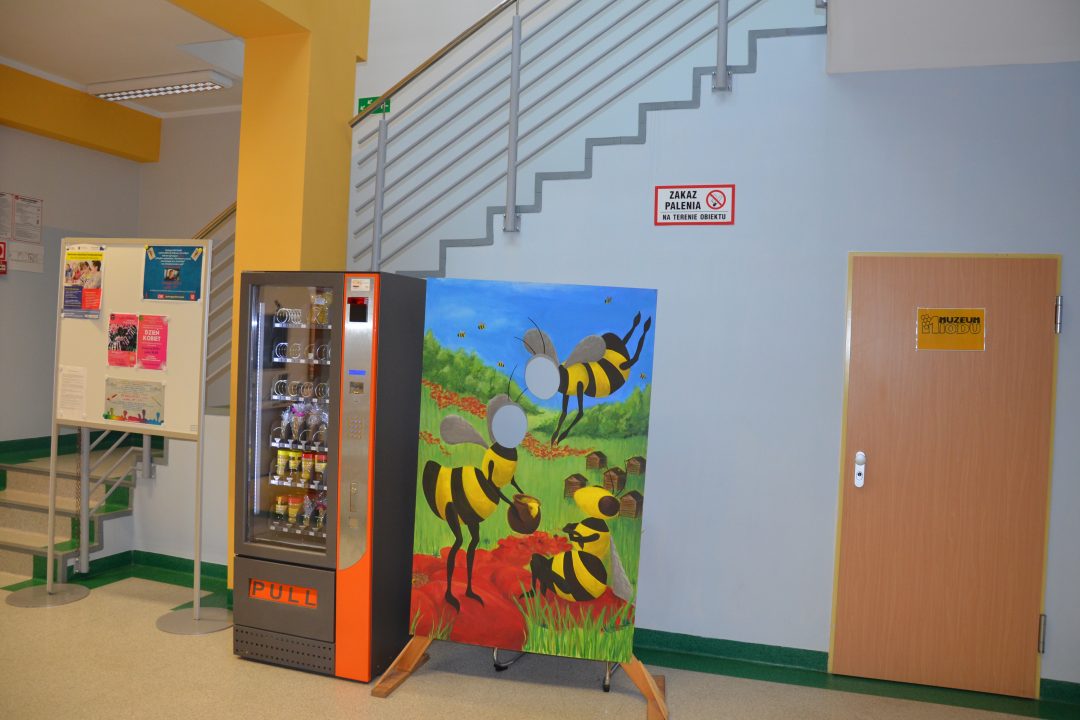 zdjęcie. korytarz biblioteki. od lewej tablica ogłoszeń, automat z przekąskami, monidło z pszczołami, drzwi do muzeum miodu.